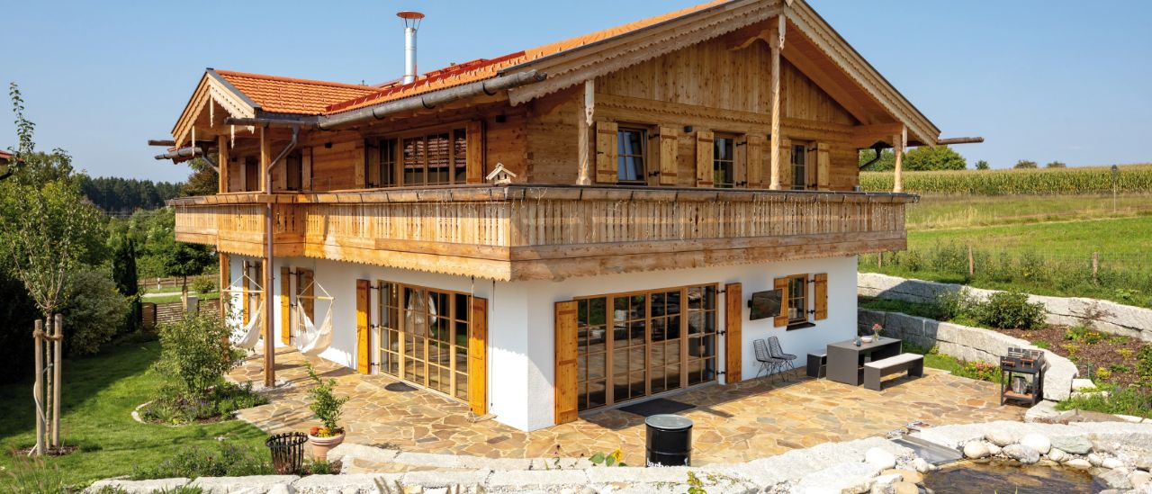 Holzhaus mit Satteldach, Balkon und vielen Fenstern, umgeben von Grünflächen und landwirtschaftlich genutzten Feldern.