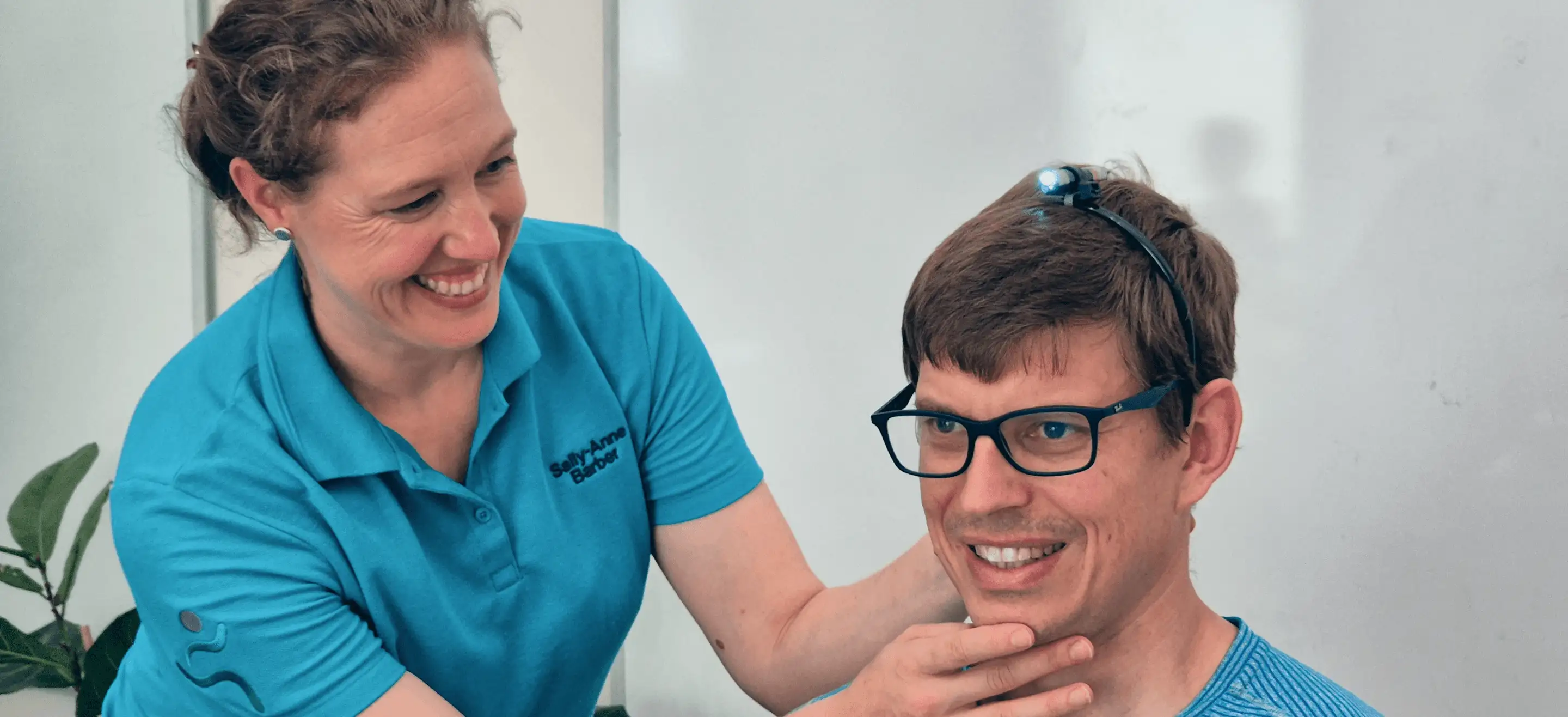 A smiling woman adjusts the chin of a young man wearing glasses, both dressed in blue shirts.