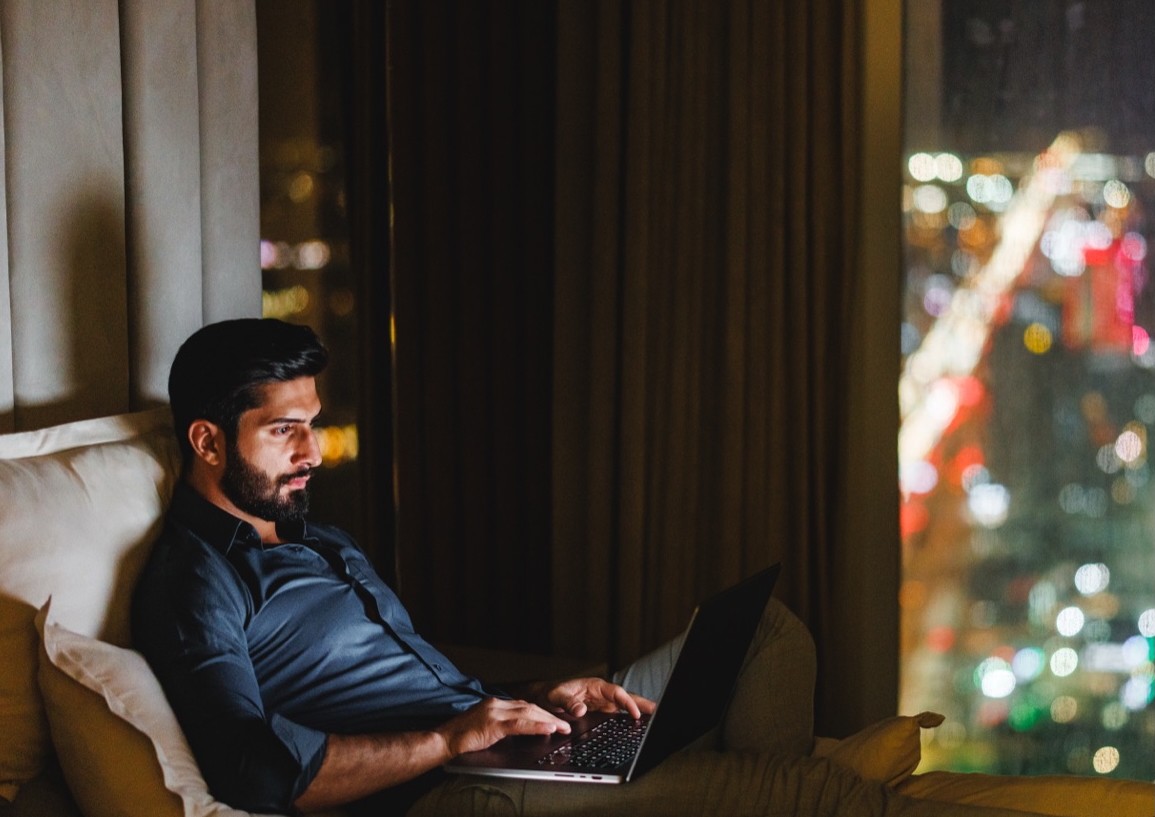 Man sitting on a sofa at night working on a laptop, with a city skyline visible through the window, representing remote management of residential buildings.