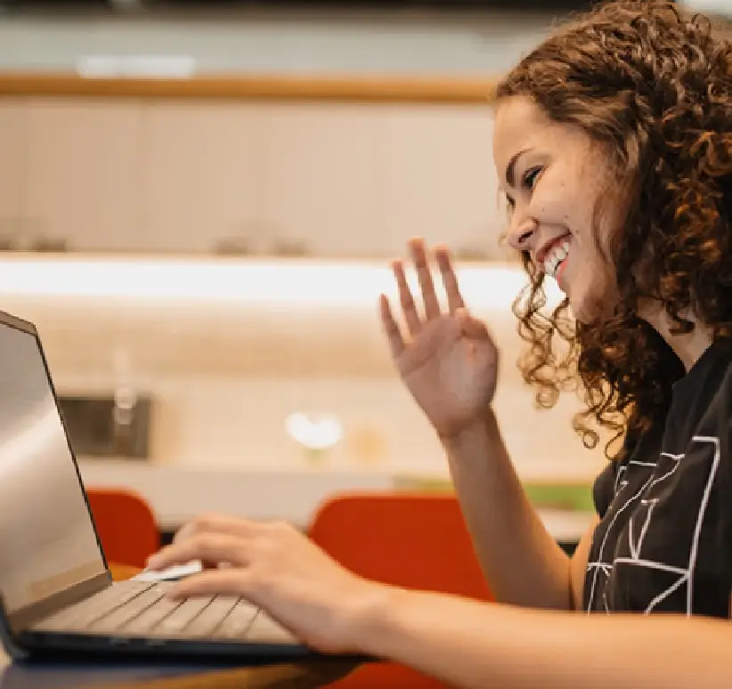 Uma mulher sentada em uma mesa, equipamentos em seu laptop, com plantas ao fundo e um interior casual e moderno.
