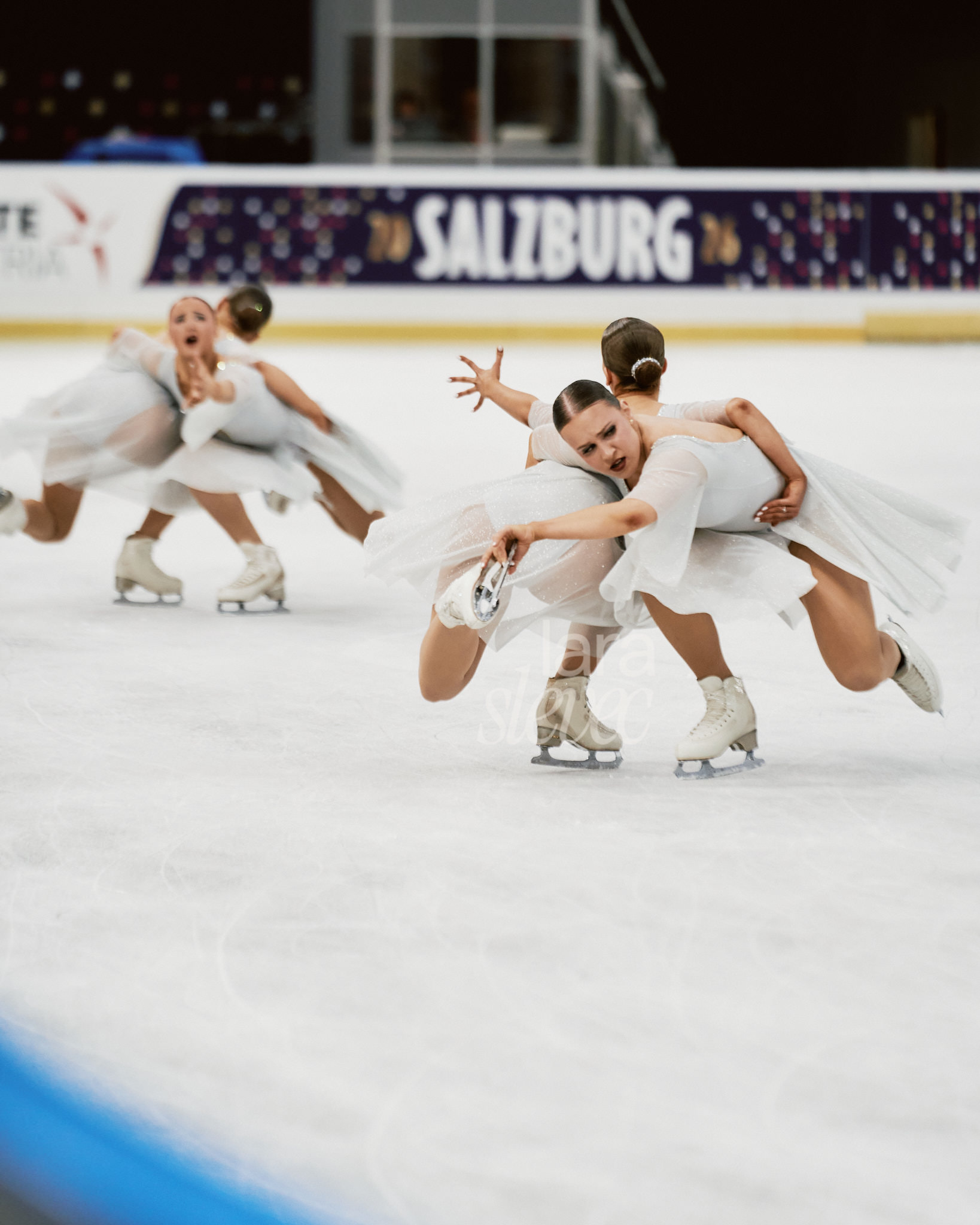 Vier Synchroneiskunstläuferinnen, die im Paar eine Sitzpiruette zeigen. Dabei haben sie die Hand zueinenader ausgestreckt und schauen sich sehnsüchtig an. Team Unique. ISU Synchronized Skating Championships 2026 in Salzburg. Fotografiert von Lara Slevec.