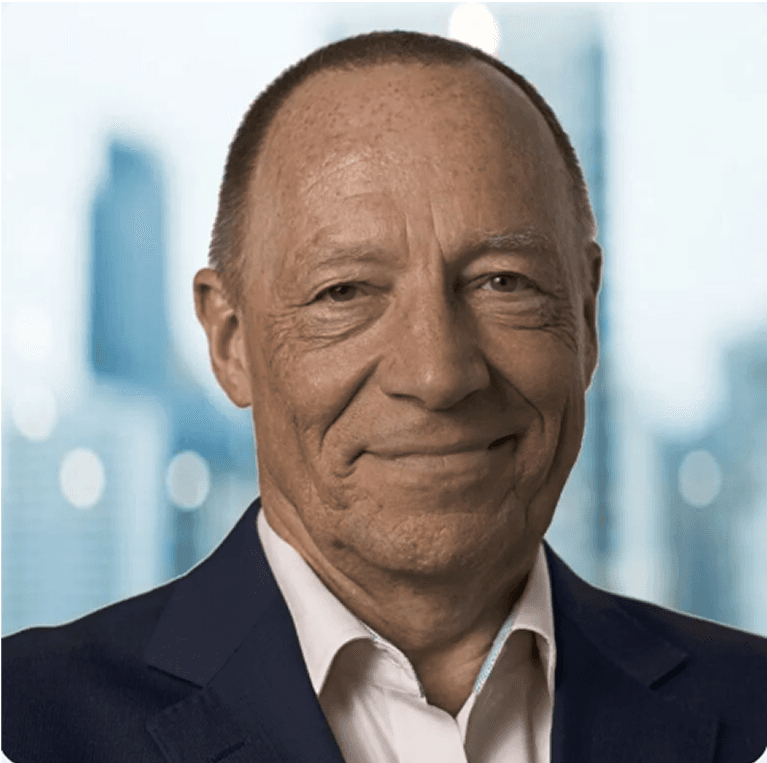 A middle-aged man with a friendly smile, wearing a dark suit, with a cityscape in the background.