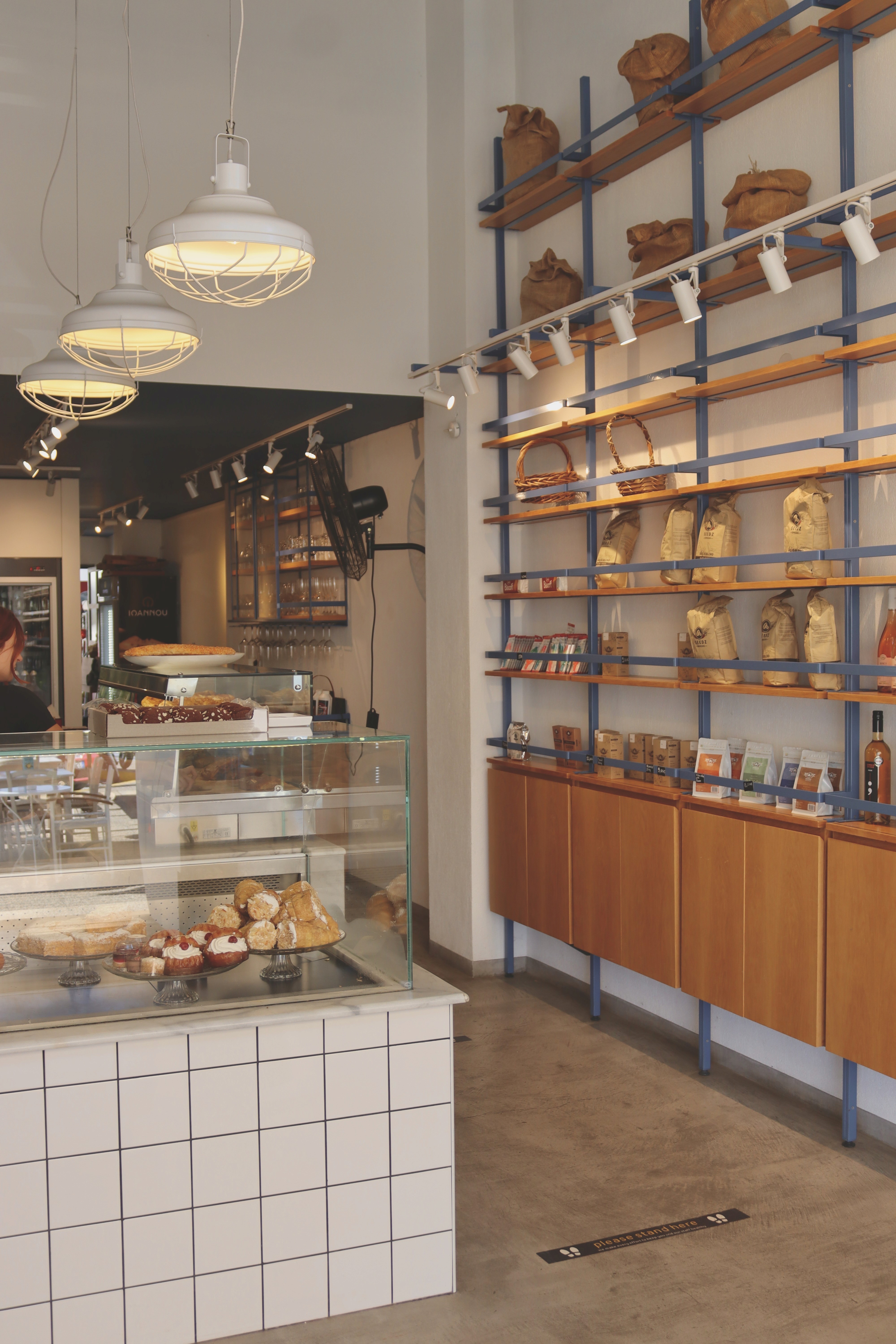 Interior de uma padaria ou bistrô com a exposição de produtos desfocada. À esquerda, uma vitrine refrigerada exibe pães e doces sobre azulejos brancos. À direita, prateleiras de madeira e metal exibem produtos embalados, com iluminação quente direcionada.