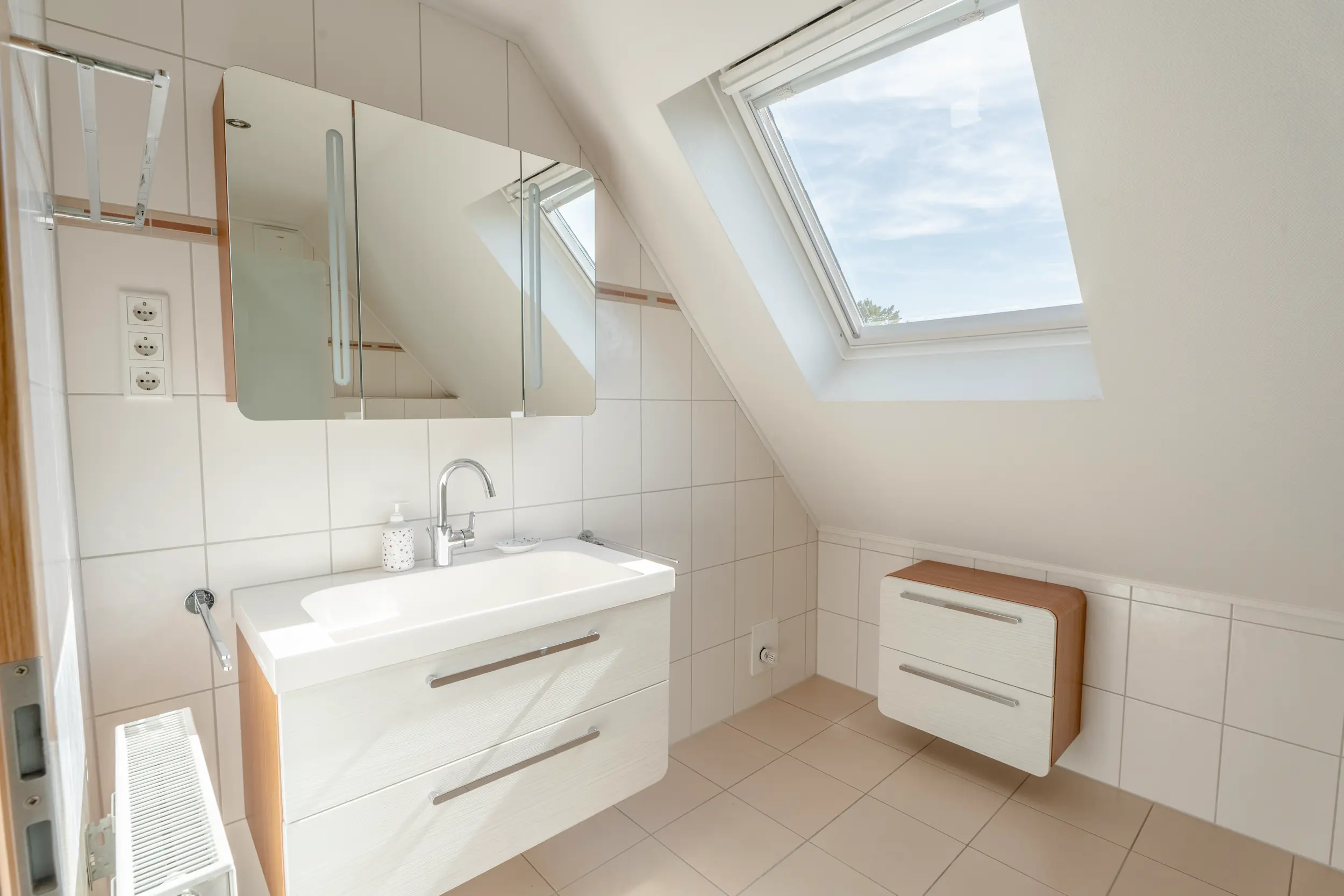 Helles Badezimmer unter einer Dachschräge mit Dachfenster, Waschbecken, Spiegelschrank und Handtuchheizkörper.