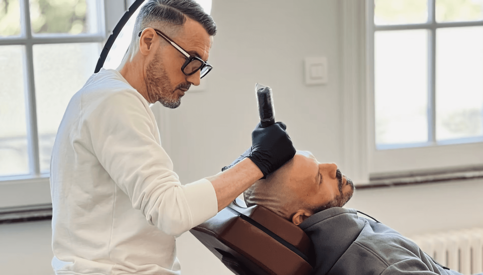 A person with glasses and gloves is tattooing on a client's arm in a well-lit studio.