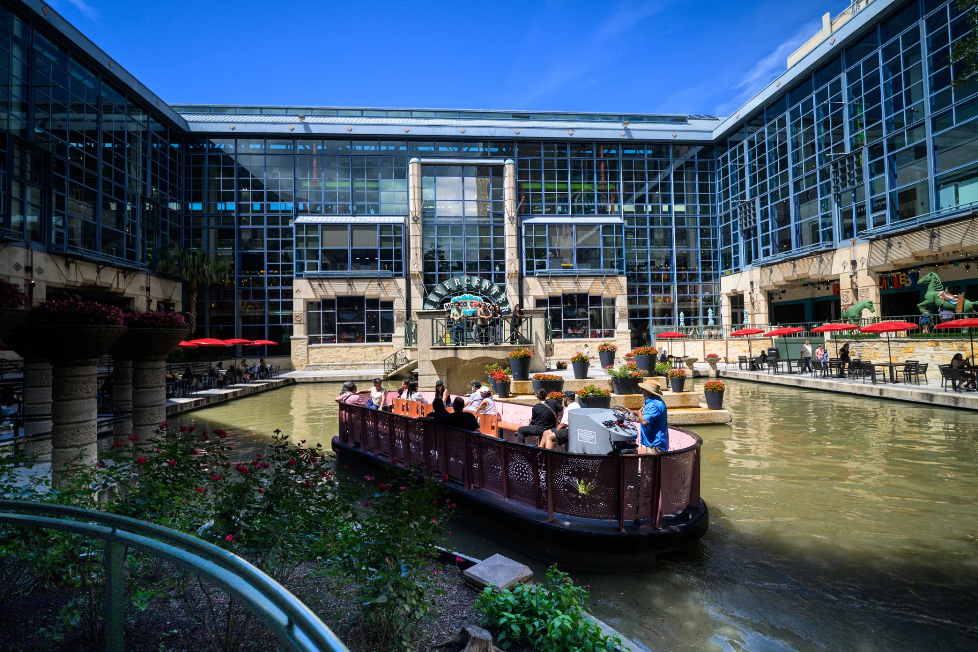 River barge with passengers on the water inside the Rivercenter courtyard, red umbrellas and the Mexico Ceaty entrance in the background