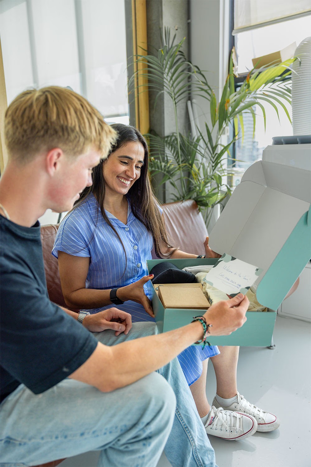 Twee mensen zitten op een bank en openen een onboarding box met een briefje en verschillende items. Ze glimlachen terwijl ze het briefje lezen.