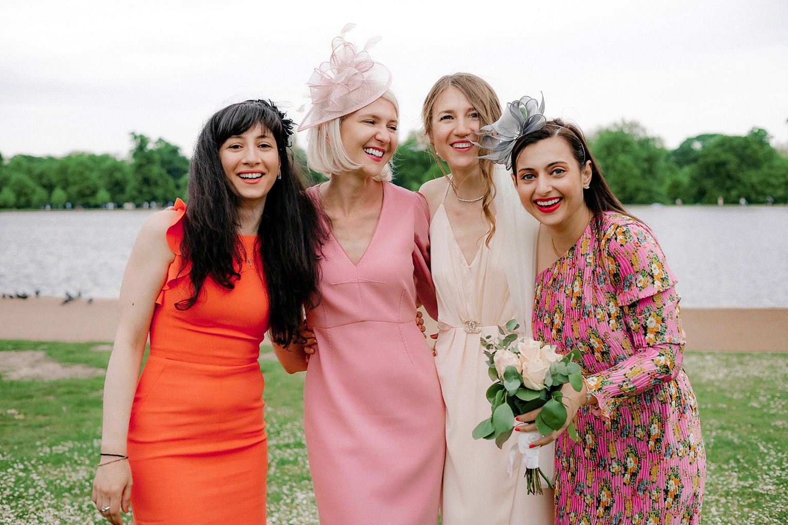Natural outdoor wedding group portrait of bride and guests smiling by the lake in colourful formal attire and fascinator hats