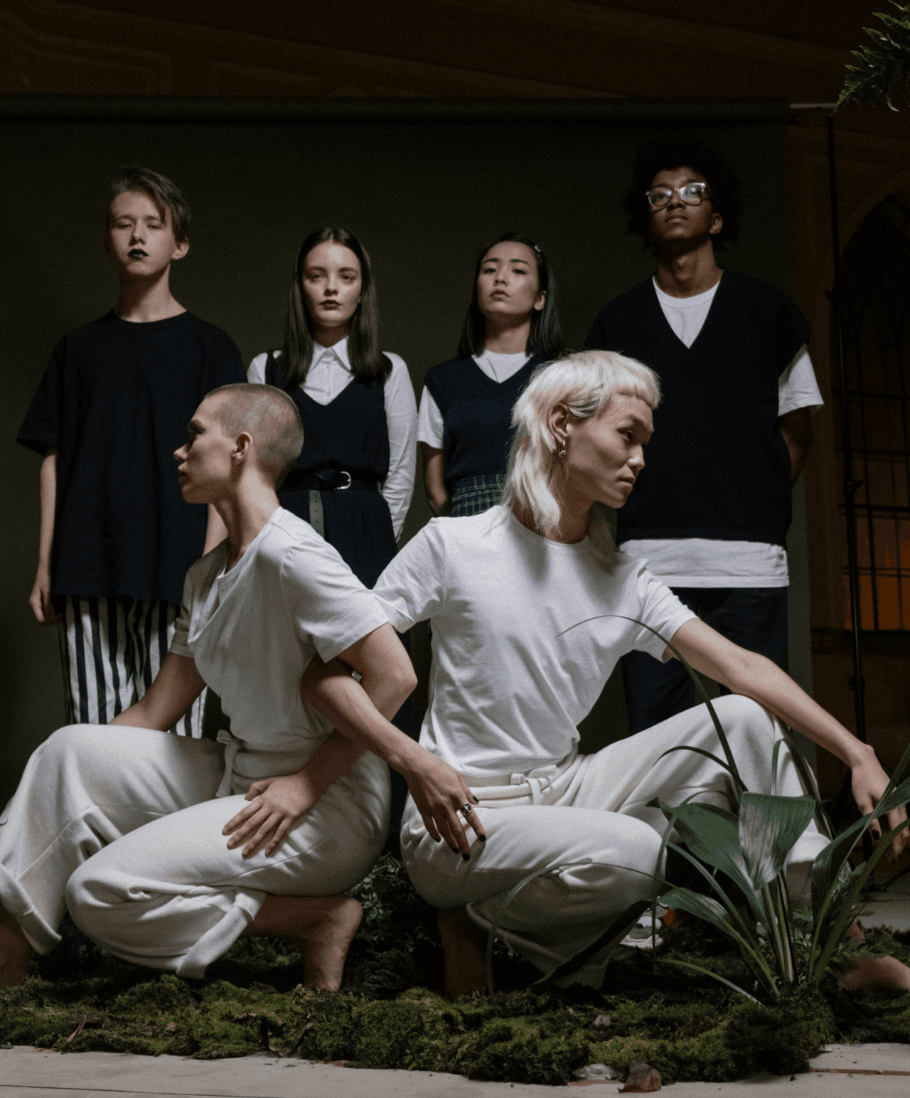 Six young people are arranged in two rows—two in white squatting in the front surrounded by moss and plants, and four standing in black attire in the background.