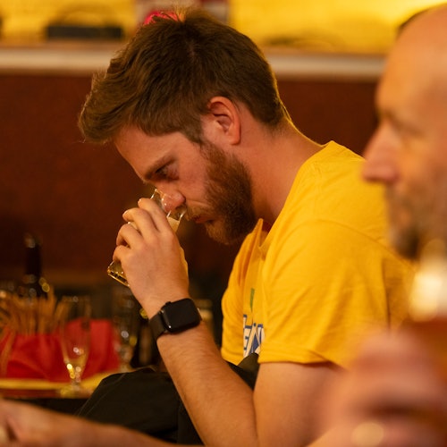 A man in a yellow shirt sniffs a drink glass while sitting indoors. Another person is partially visible in the foreground.