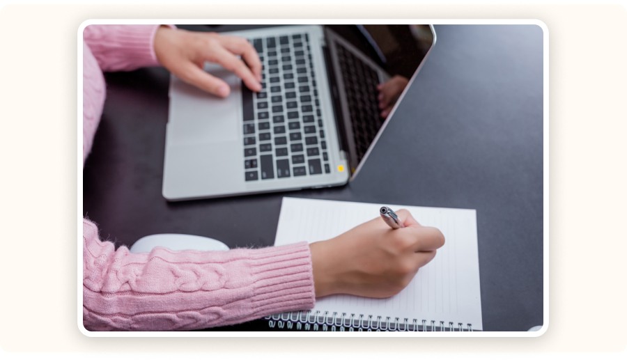 Person using laptop while writing notes in notebook on desk