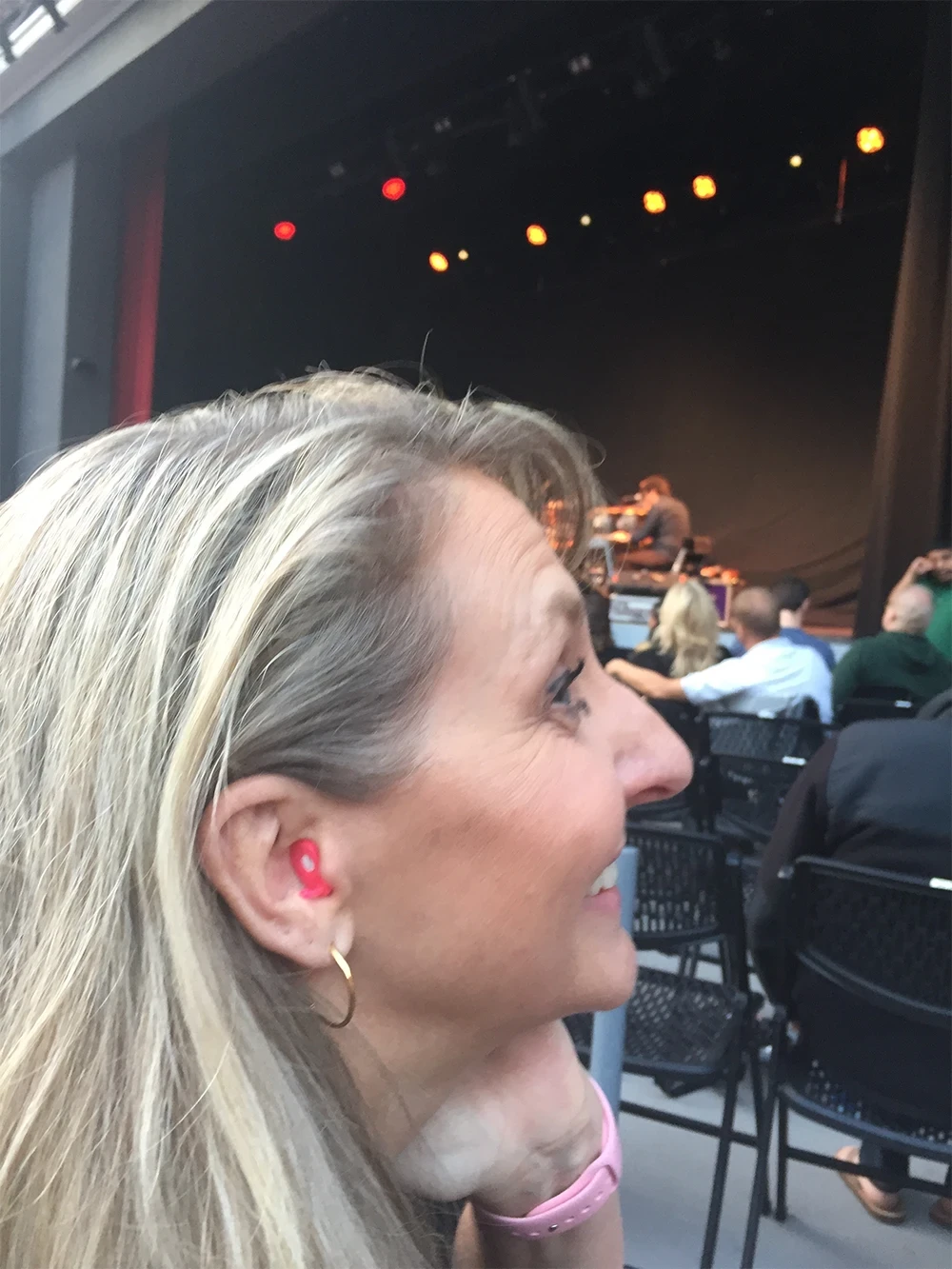 A woman with long blonde hair sits in a theater with dim lighting and blurred audience in the background.