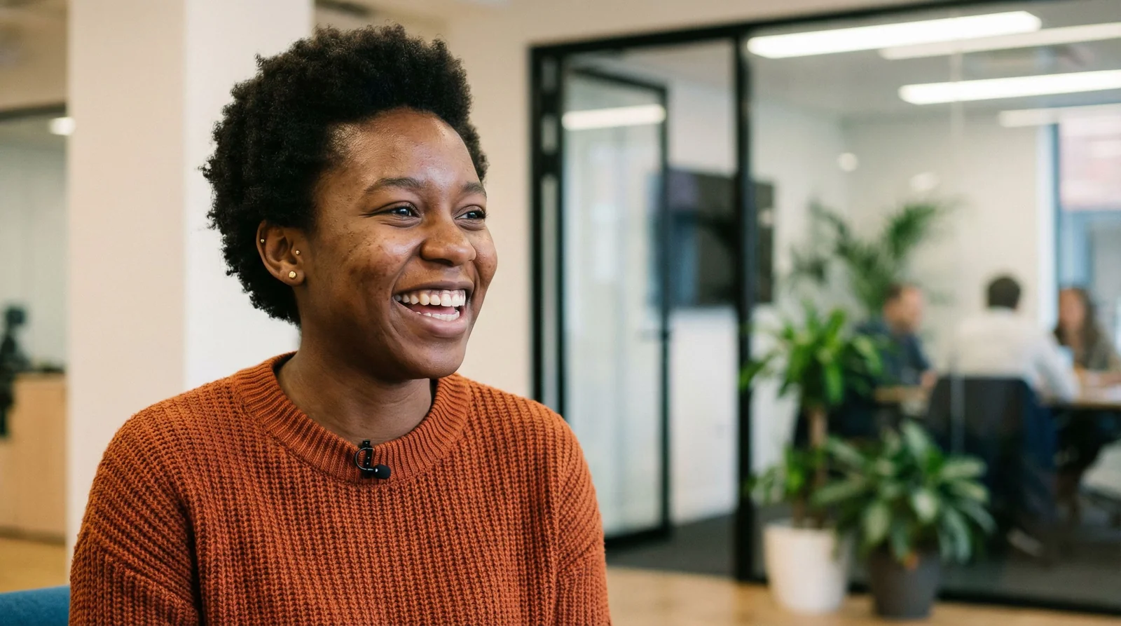 Professional in burnt orange sweater mid-laugh, lavalier mic clipped to collar, modern office with glass walls and plants in bokeh