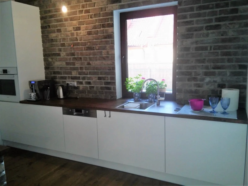 Nowoczesna kuchnia na wymiar z białymi frontami, ciemnym blatem i oknem, zamontowana na tle ściany z czerwonej cegły.