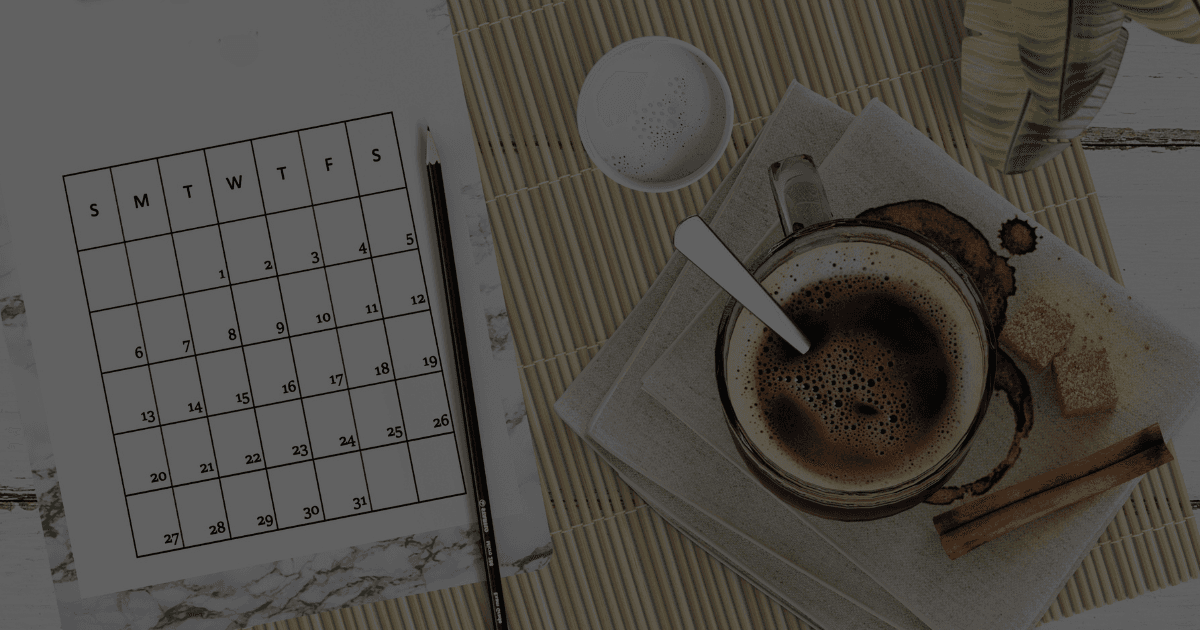 A top view of a desk with a calendar, a coffee mug filled with a foamy liquid and a few snacks and cinnamon sticks sit beside.