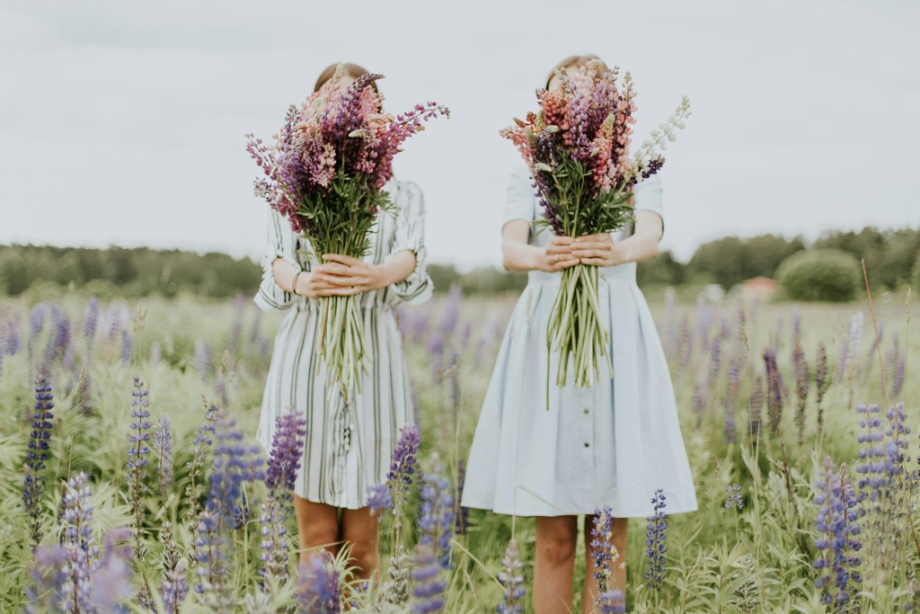 Two women in summer dresses stand in a blooming field of purple and pink lupines, each holding large bouquets of wildflowers that cover their faces, creating a serene and whimsical scene.