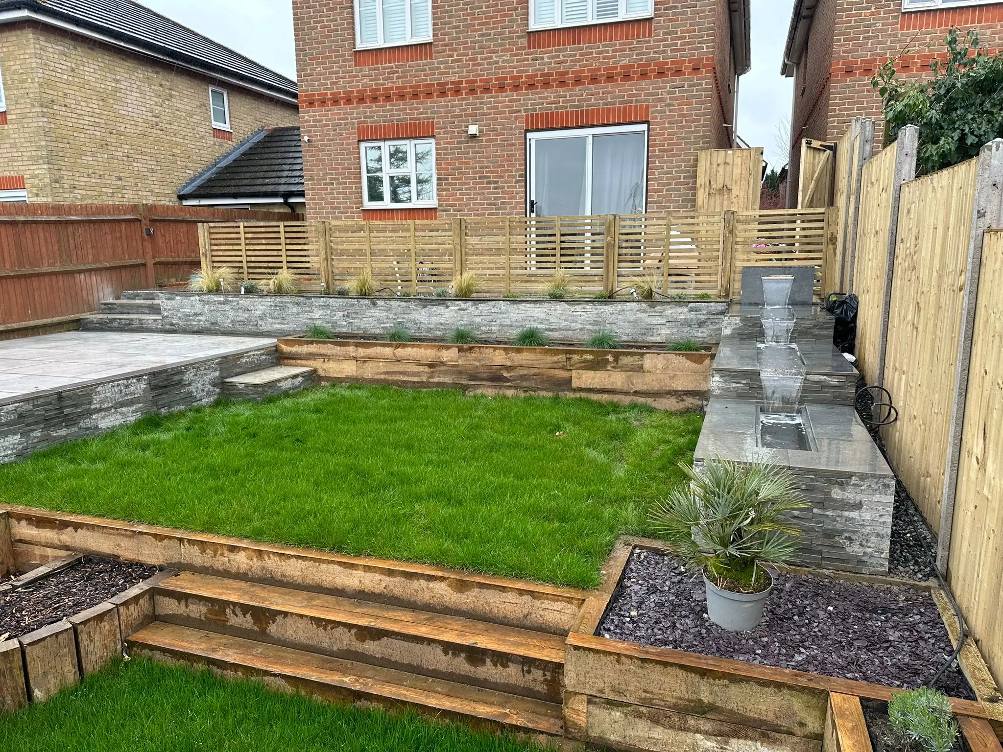 A backyard garden featuring grass, wooden borders, and stone planters next to a brick house.