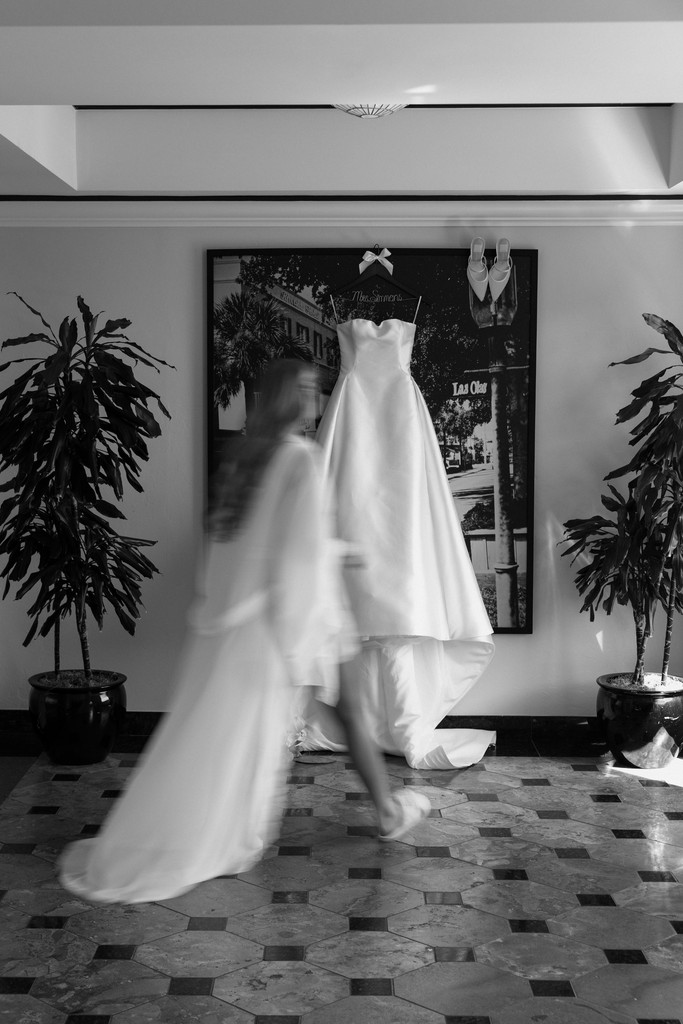 Wedding photographer in South Florida captures a bride walking across the room in her robe with her wedding dress hanging on the wall.
