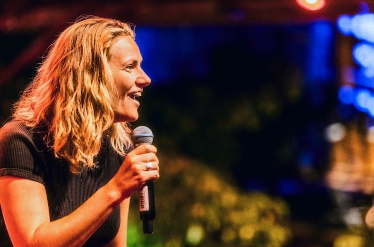 A woman with wavy hair smiles while holding a microphone, speaking into the audience against a colorful backdrop.