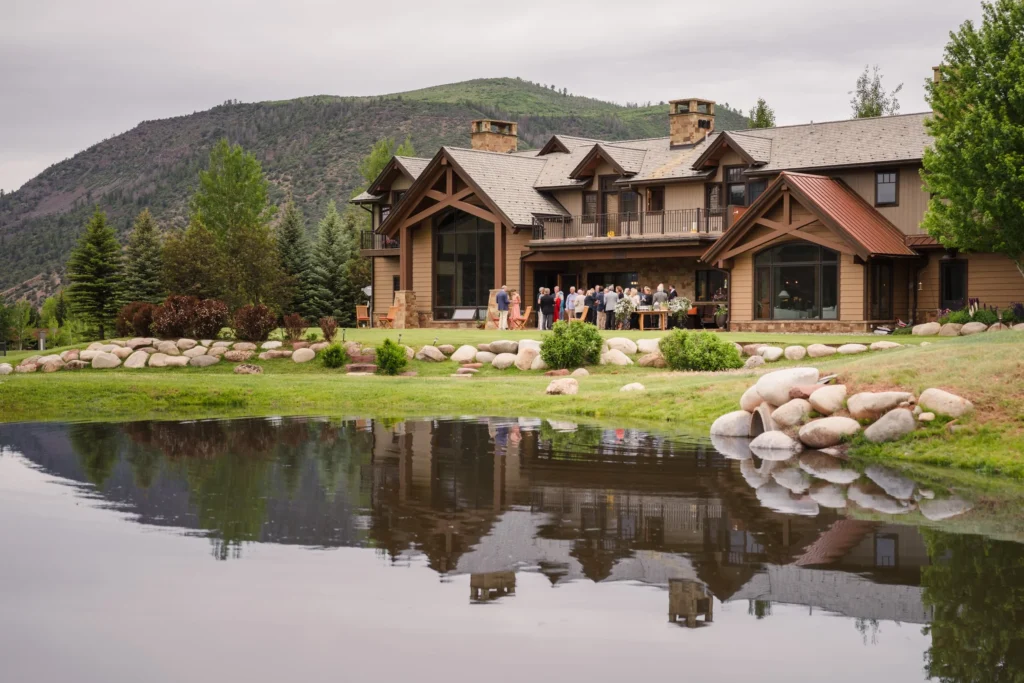 A wide landscape shot showing a large, rustic timber-framed lodge reflected in a calm pond during a Chaparral Ranch Wedding.