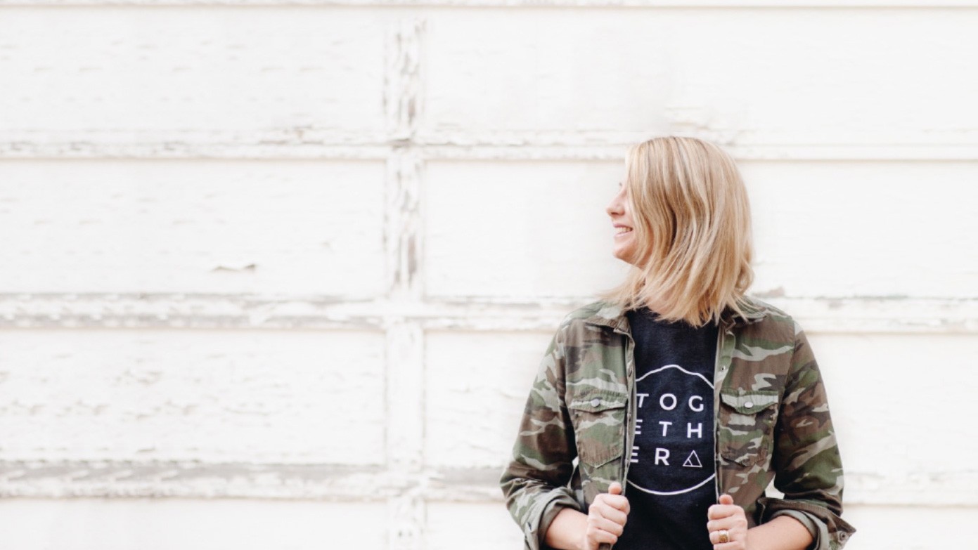 Blonde woman in camo jacket smiling against white background.