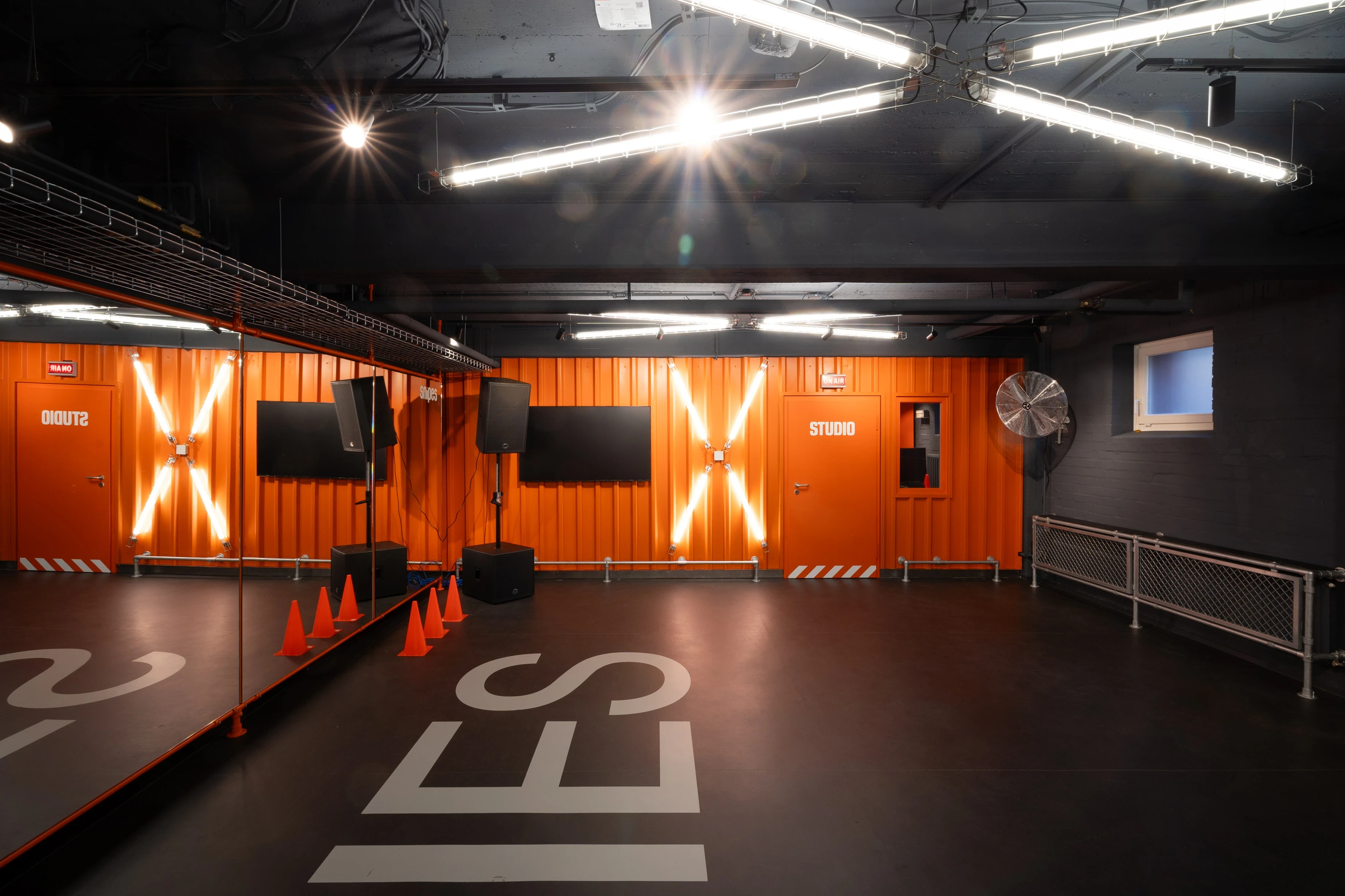 Dance room at SNIPES BASEMENT with mirrored wall, orange container-style studio doors with illuminated X marks, wall screens, speakers and traffic cones on a black floor.