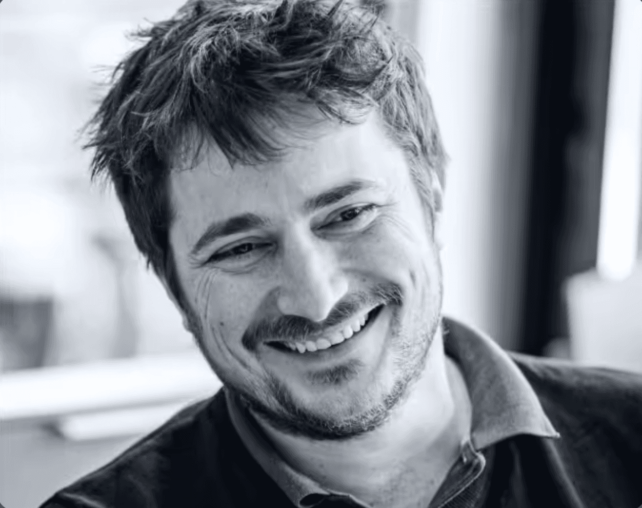 A man with glasses and a beard is looking directly at the camera, seated in a cozy, dimly lit café.