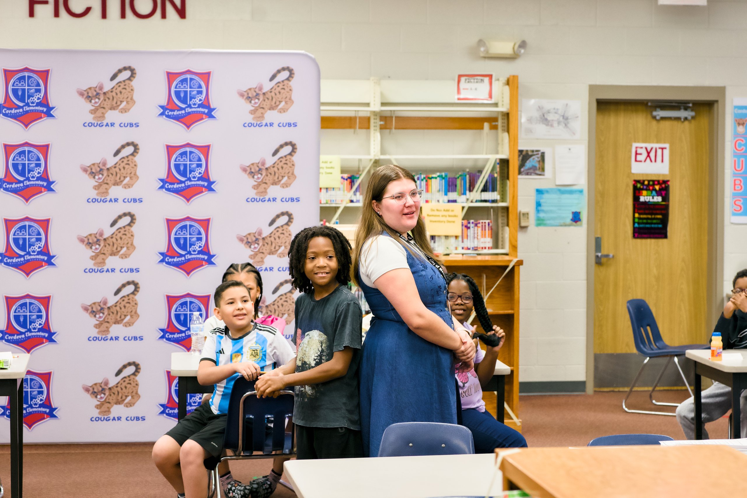 Children and a teacher pose in front of a Cougar Cubs backdrop in a school library