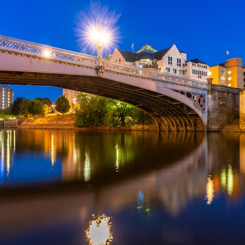黄昏时分,灯火辉煌的桥横跨在宁静的河面上,河水倒映着建筑和树木的剪影。