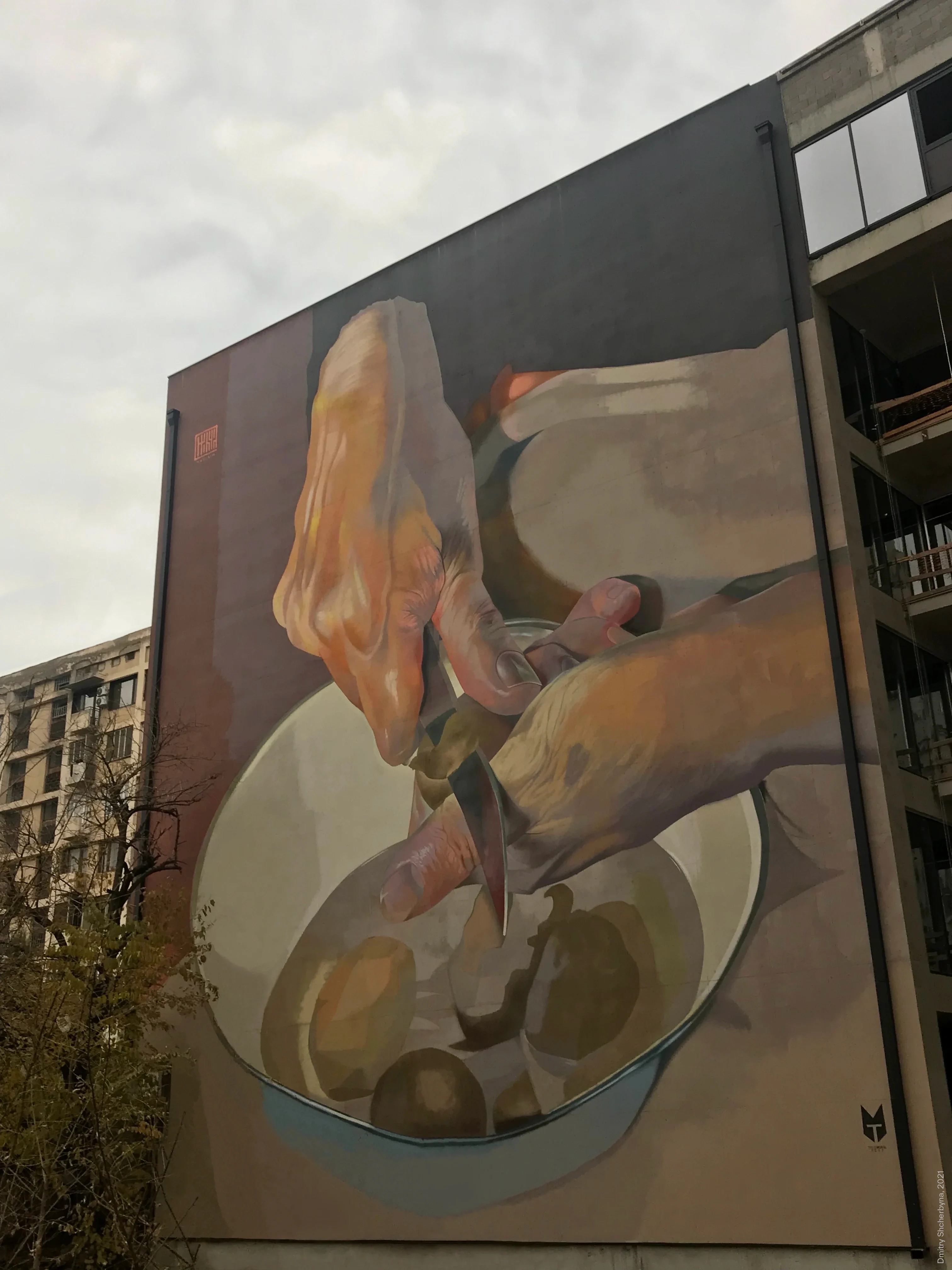 A photorealistic mural on the side of a building, showing a close-up of two hands peeling potatoes with a knife over a bowl, rendered in warm, earthy tones.