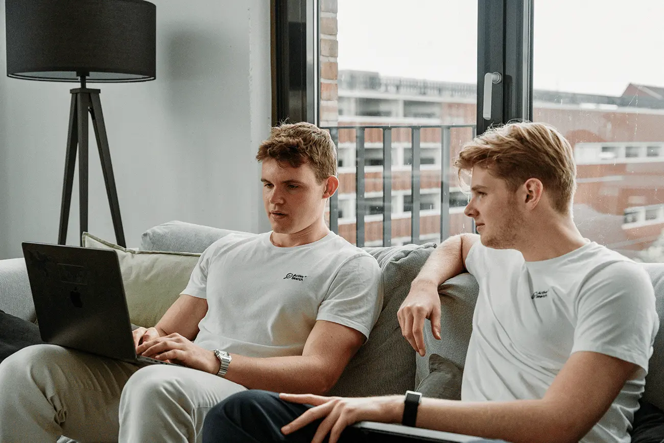 Two women are standing side by side against a beige wall, smiling confidently, one wearing a green shirt and the other in a dark blazer, with soft lighting casting gentle shadows.