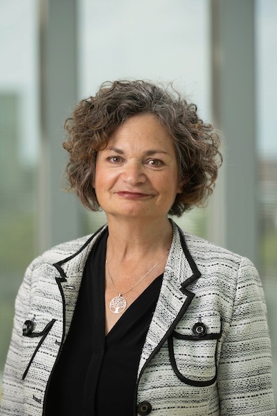 A woman with curly hair smiles, wearing a black top and a white patterned blazer, standing in a bright indoor setting.