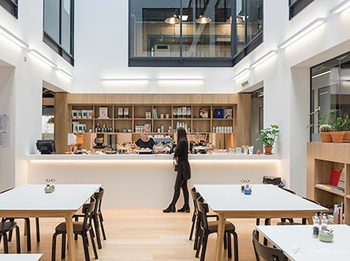 A bright, modern café-style workspace with white tables, wooden flooring, and a counter where one person serves another.