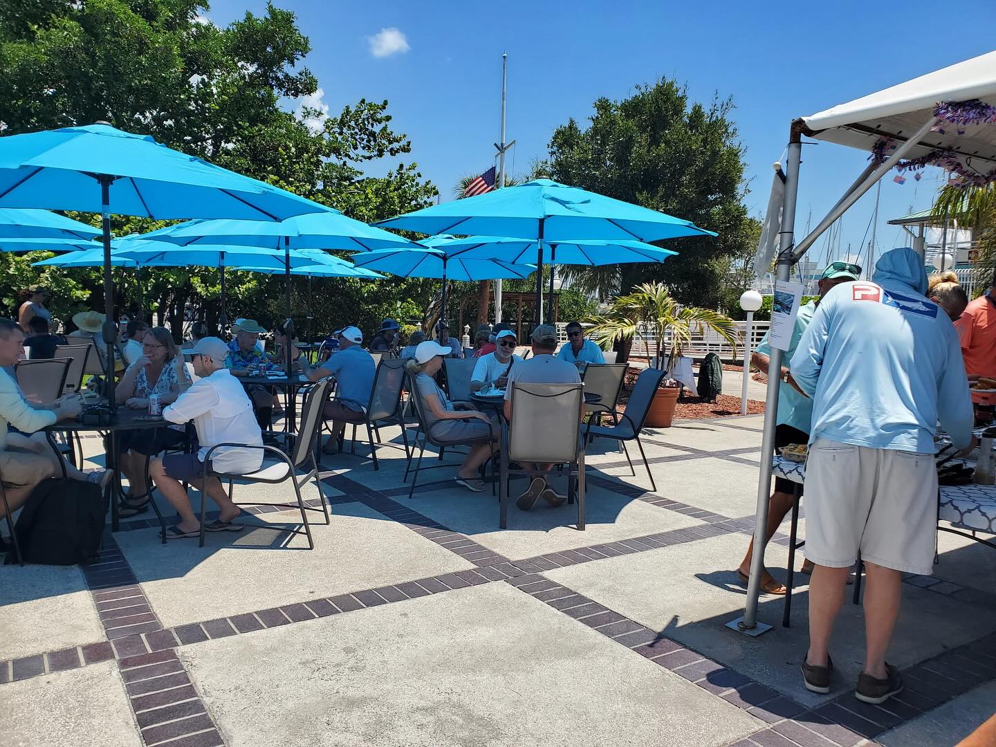 Outdoor café with people seated under vibrant blue umbrellas enjoying a sunny day, surrounded by lush greenery, with an American flag visible in the background.