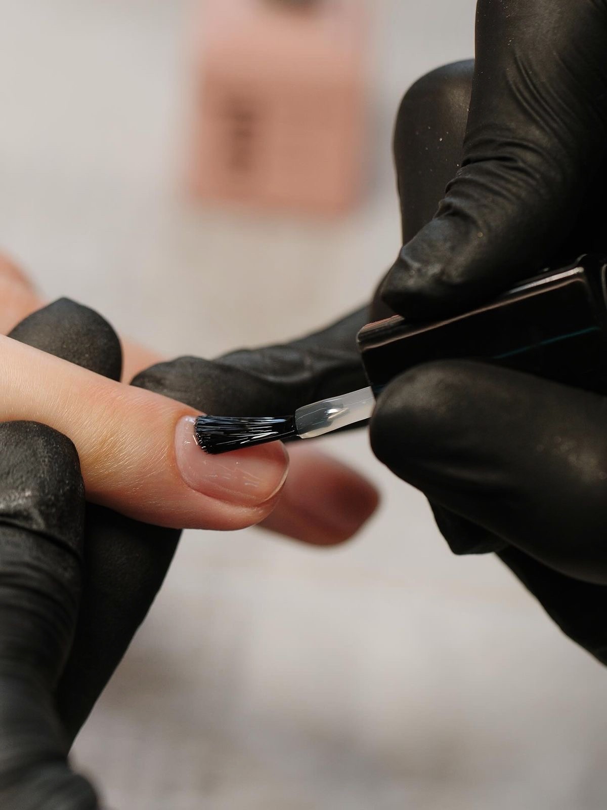 Professional nail technician wearing black gloves applying a clear top coat or gel polish to a client's fingernail during a luxury manicure salon service.