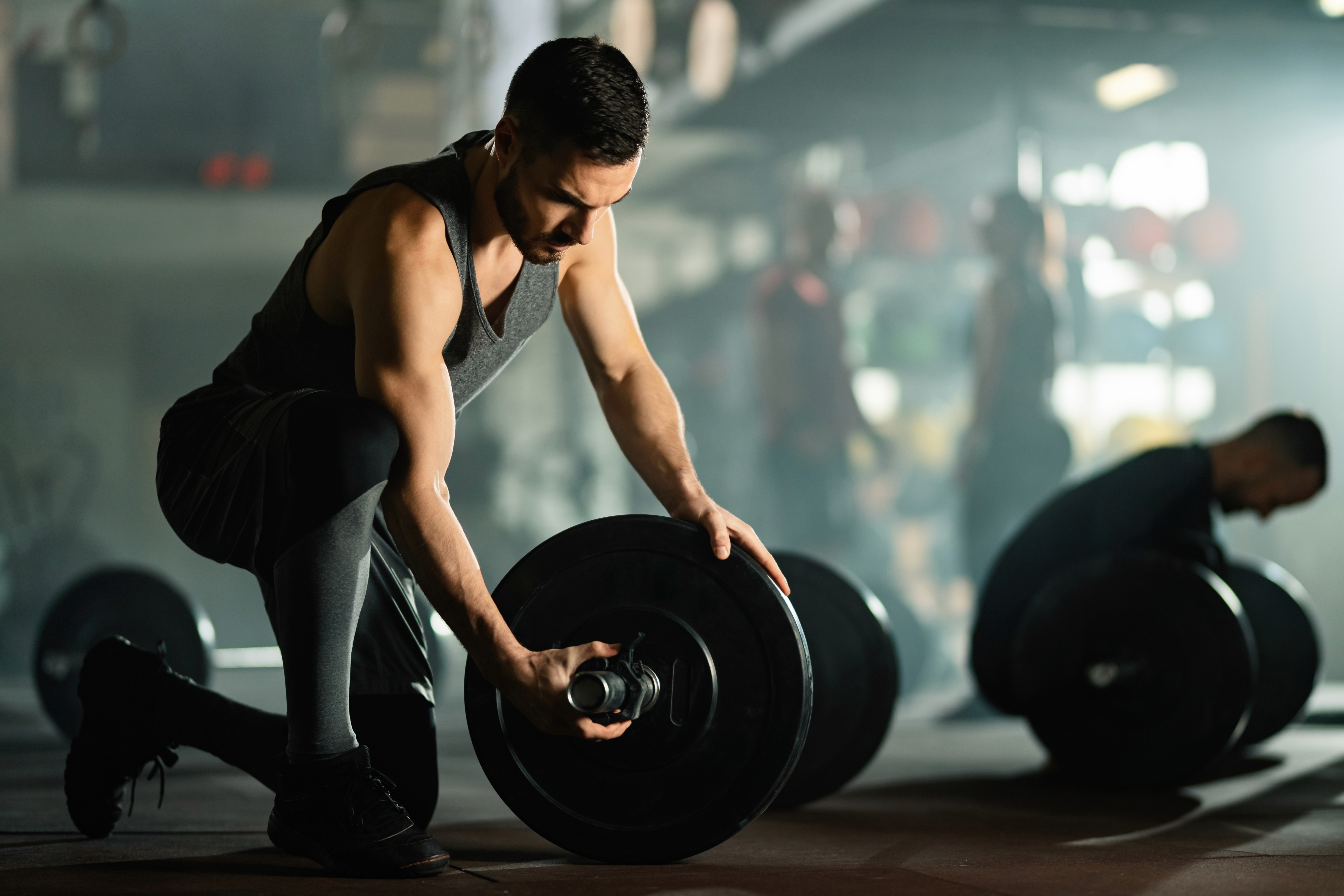 A young man in a gym kneeling down while adjusting the weight plates on a barbell. He is wearing a sleeveless gray shirt and appears focused on his task. The gym environment is dimly lit, with a slight haze in the background, giving a dramatic effect to the scene. There are other people in the background, some also lifting weights, but they are out of focus, adding depth to the image. The atmosphere conveys a sense of concentration and dedication during a weight training session.