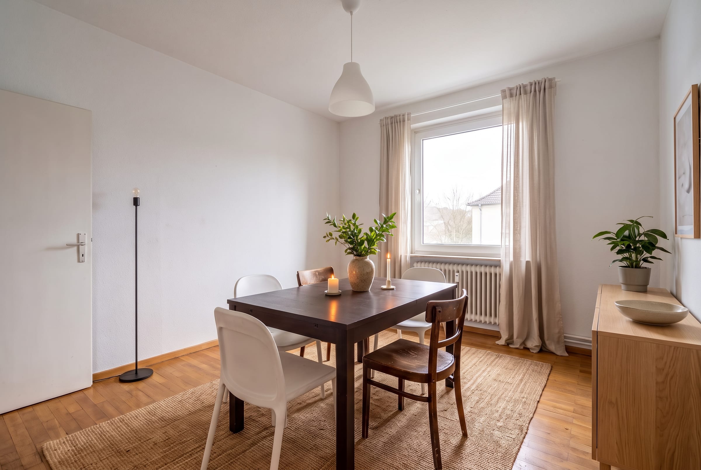 Kleiner dunkler Holztisch mit weißen Schalenstühlen und Holzstühlen mit Sprossenlehne auf einem Juteteppich, daneben ein Holzsideboard und Grünpflanzen