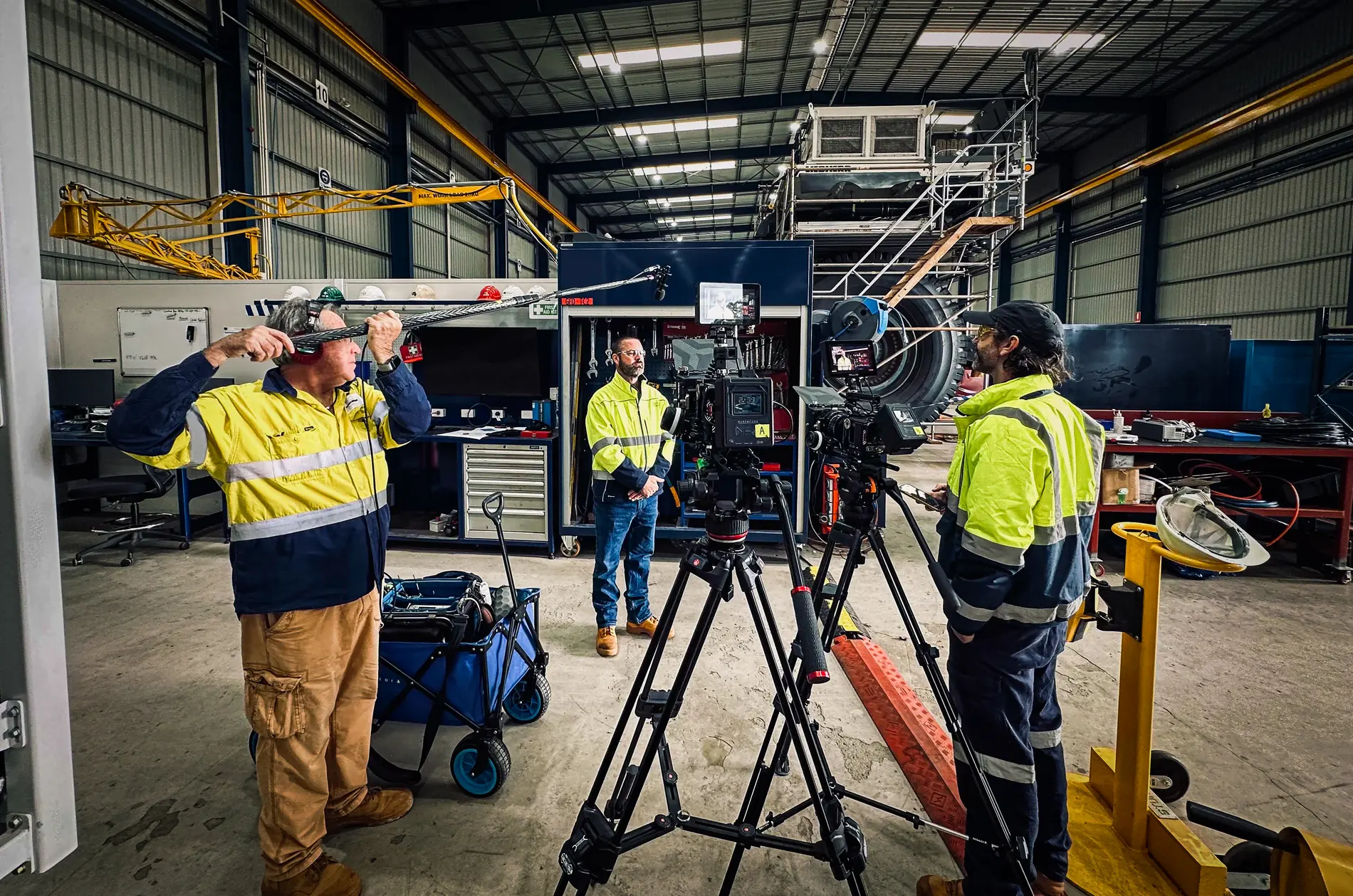Film crew with cameras on tripods recording a worker in high-visibility clothing inside a workshop