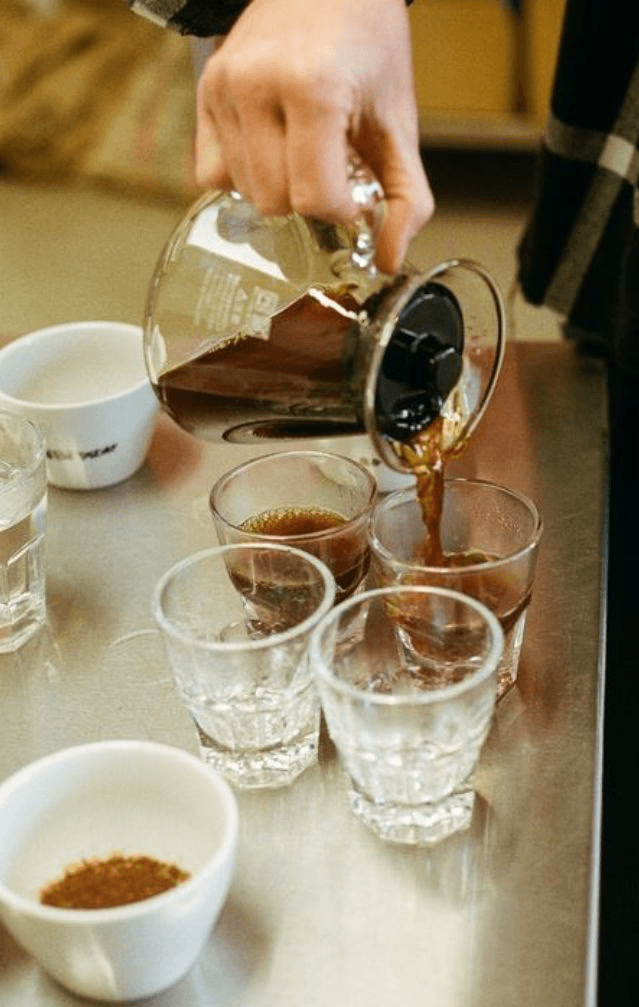 Cozy cafe corner with a round terrazzo table surrounded by wooden chairs, decorated with potted plants and a large mirror reflecting warm light.