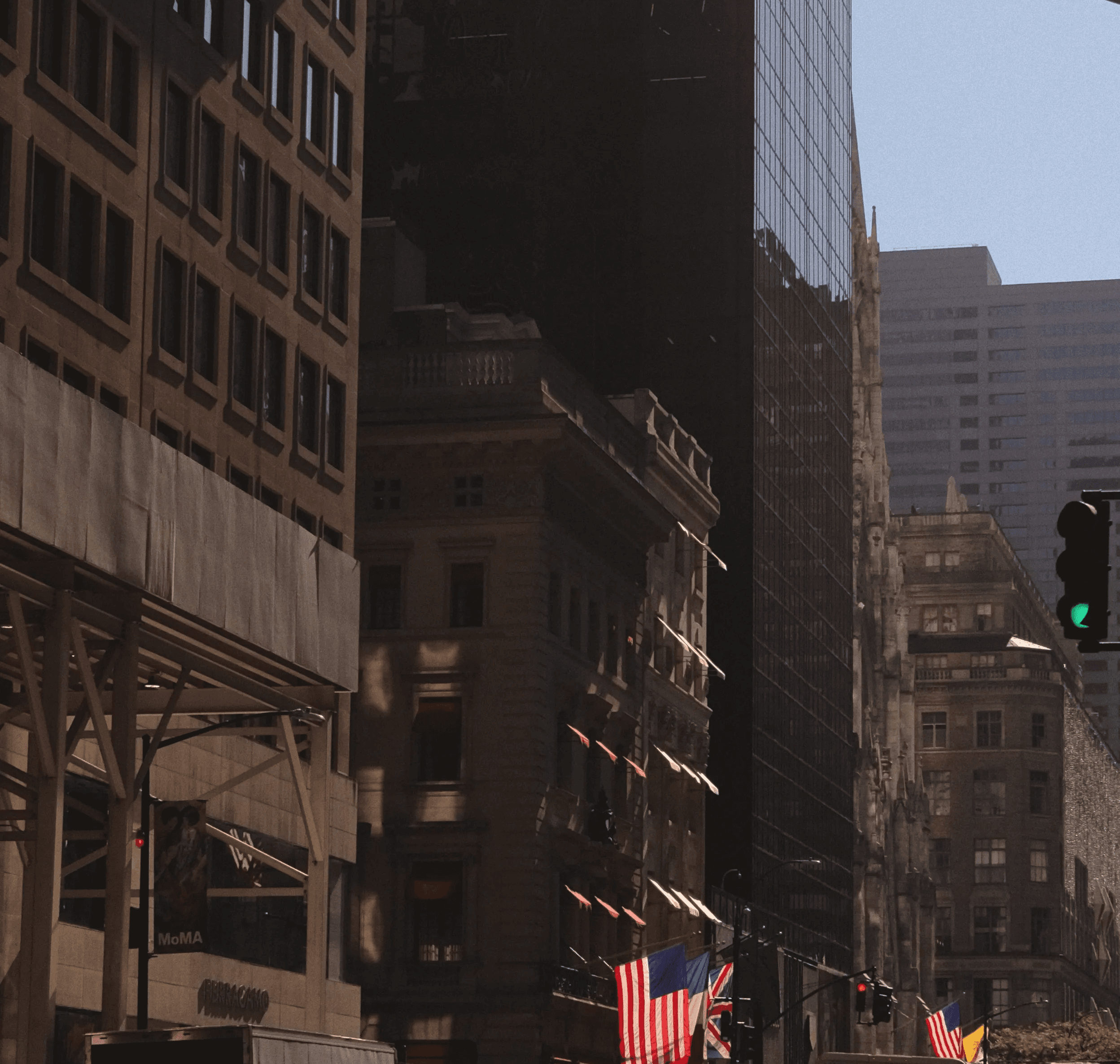 Street-level view of Midtown Manhattan near MoMA, with yellow taxis, American flags and a pedestrian crossing a sunlit intersection.