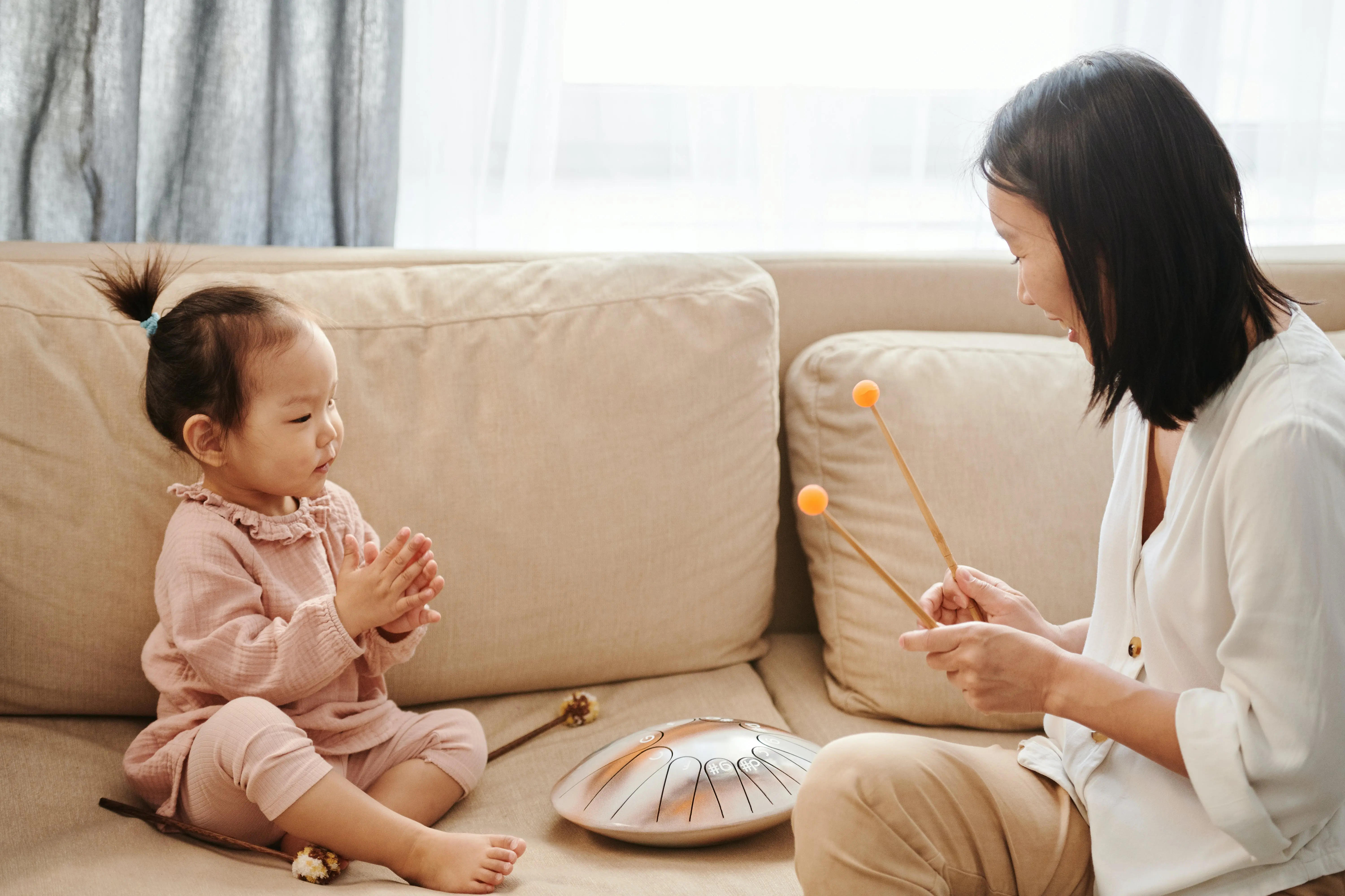 Mother playing a tongue drum instrument with her toddler on a sofa, encouraging creative play and bonding time.