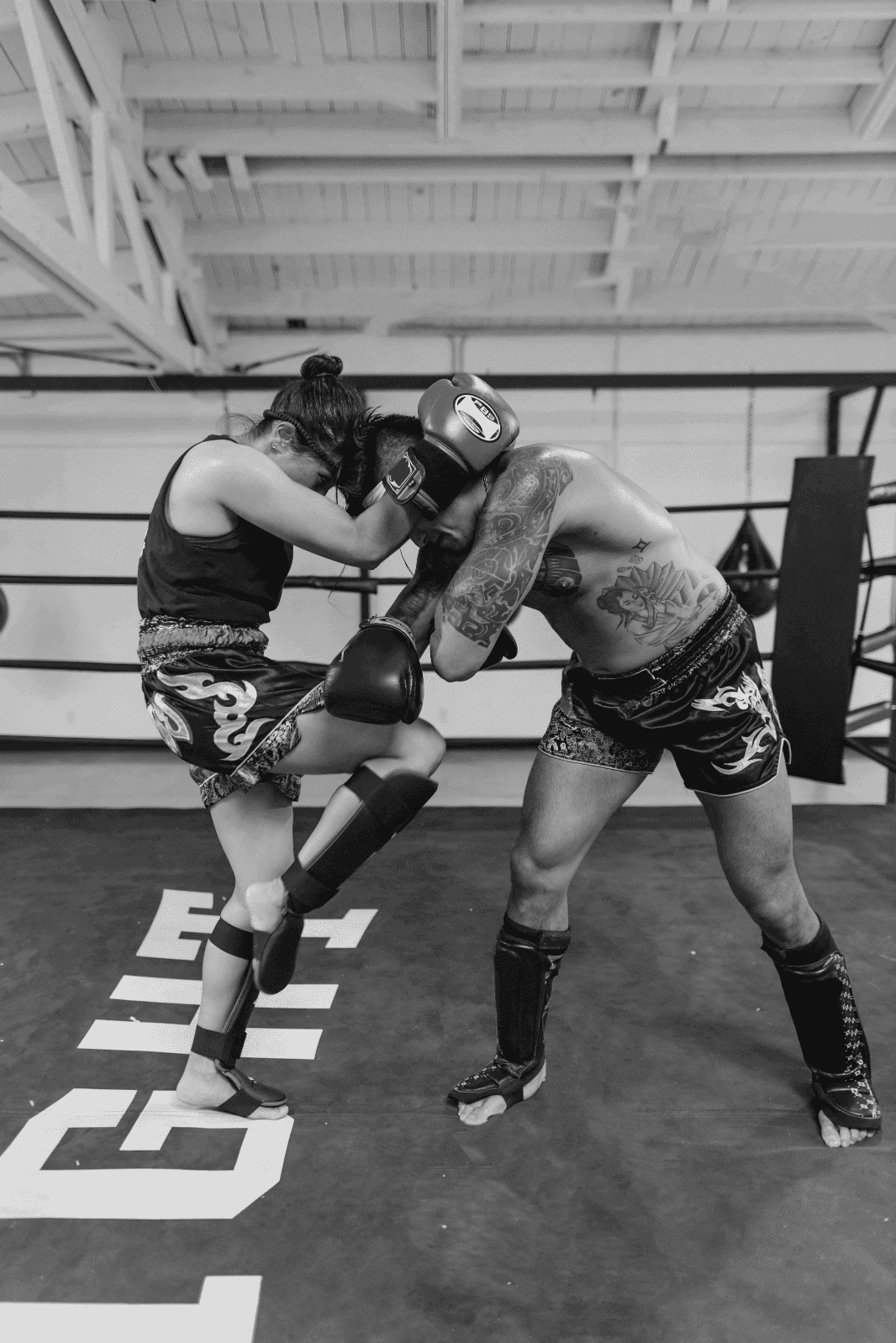 A Muay Thai training session where a female fighter delivers a knee strike from the clinch against a male training partner inside a boxing gym.