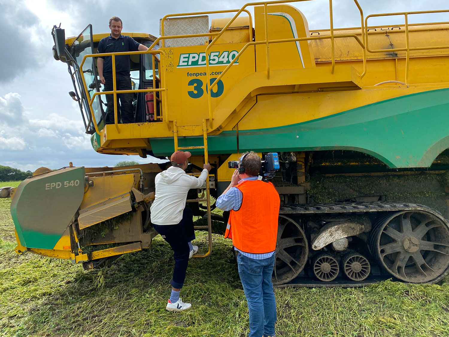 Joe climbing the ladder into the cab of a pea harvester. There is a camera operator wearing an orange hi-vis vest.