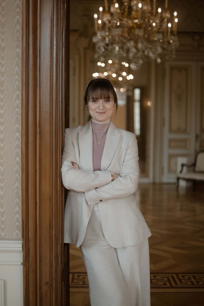 Hochzeitsplanerin Emily steht angelehnt in einem Wiener Ballsaal und schaut lächelnd in die Kamera. 