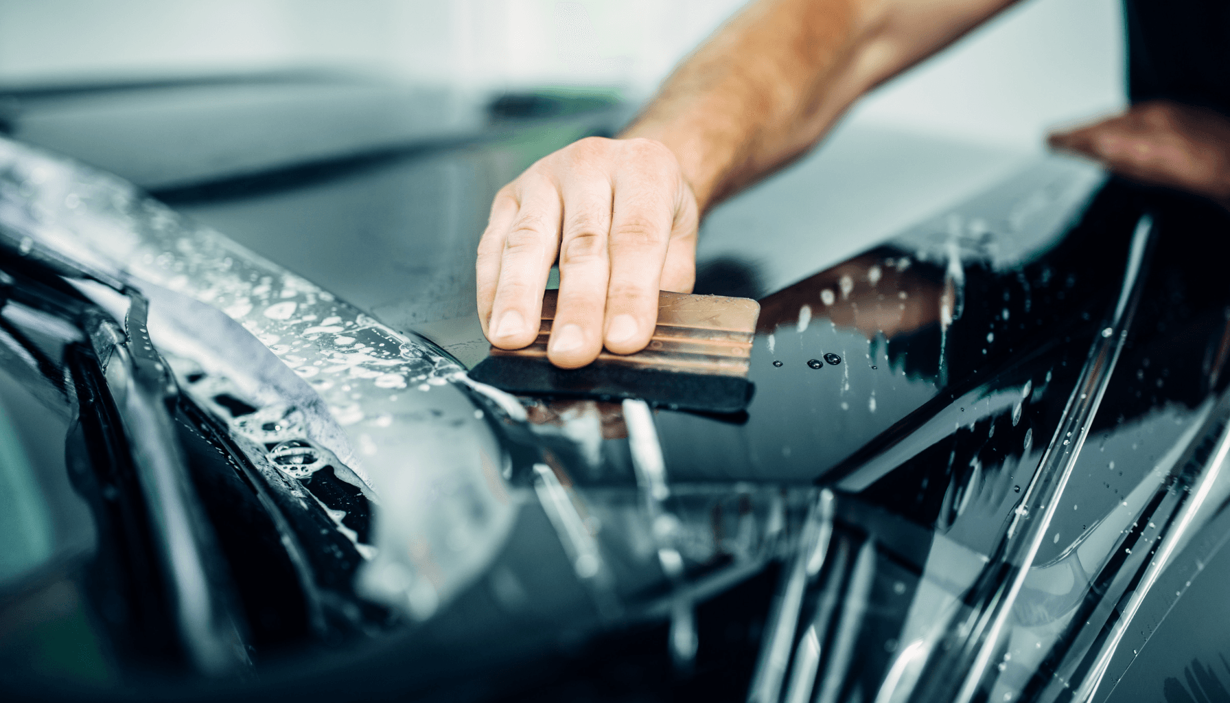 Das Bild zeigt eine Hand, die mit einem Schaber eine Autolackierung reinigt.