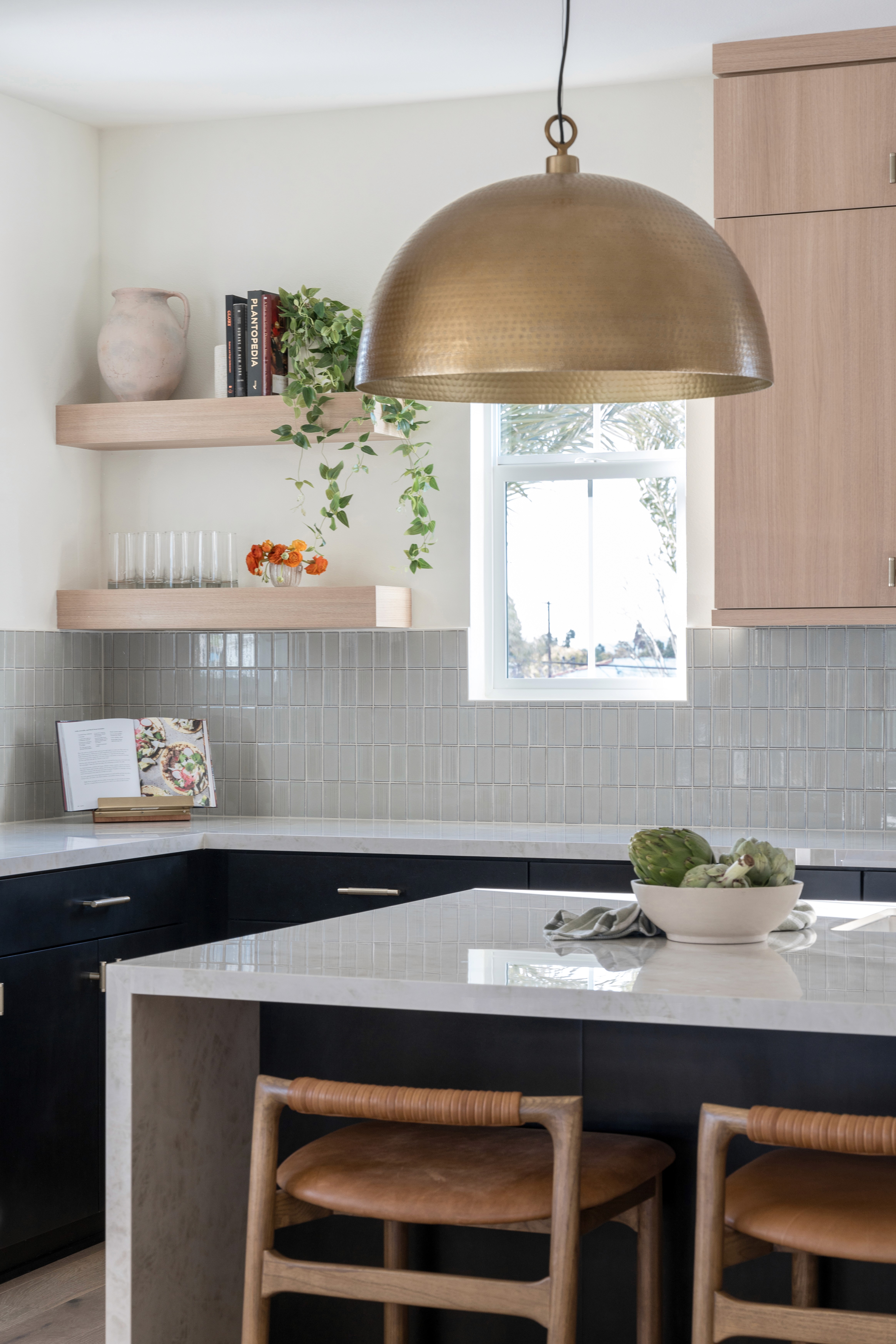 Kitchen island with large, brass pendant lighting, woven counter stools, light wood upper cabinets, sea green tile backsplash, and black lower cabinets