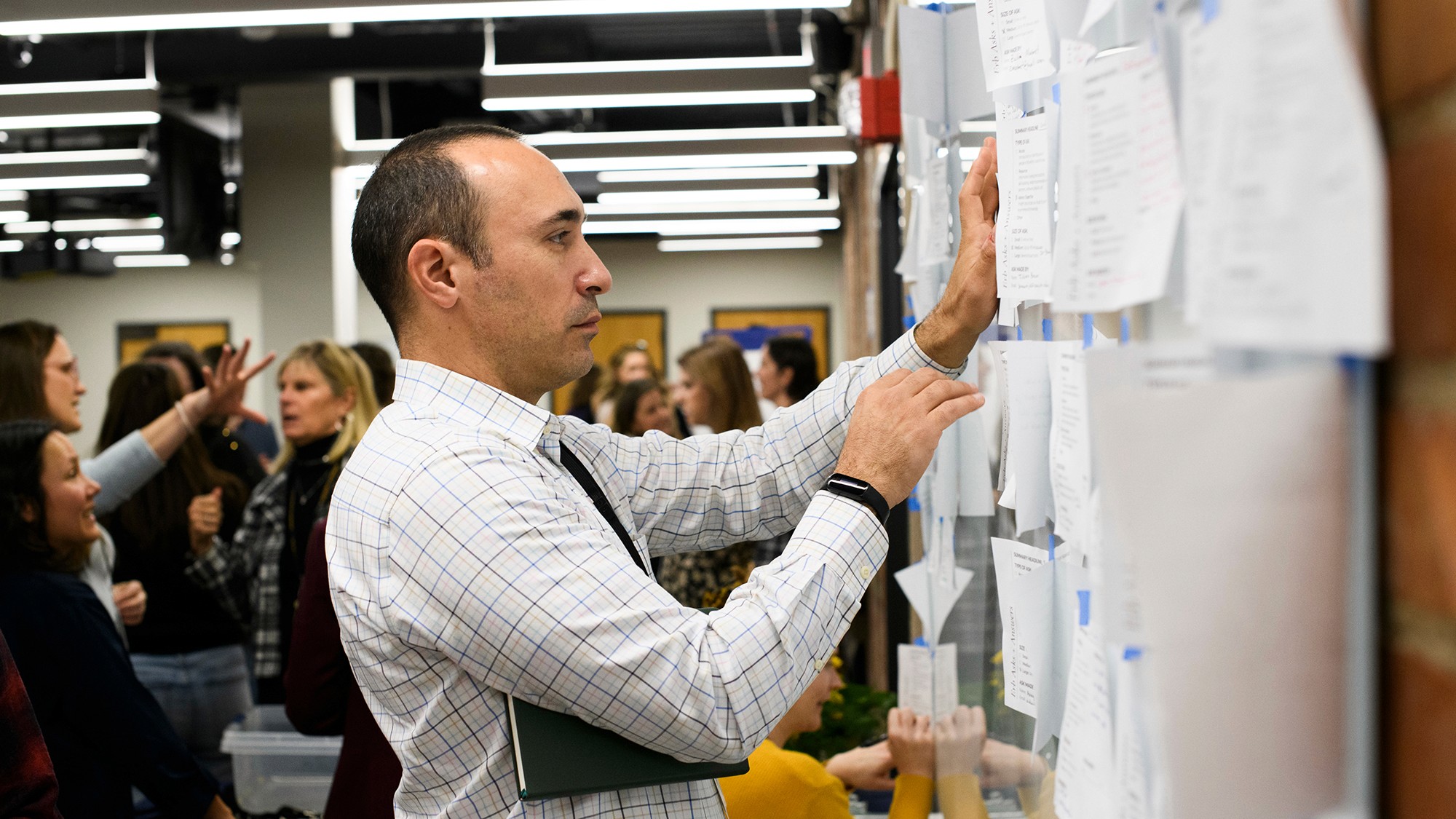 A person in a white shirt pins documents to a wall in a busy workspace, engaged in collaborative discussion.