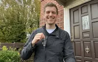 Man in grey jacket smiling and holding up house keys in front of a brick house entrance
