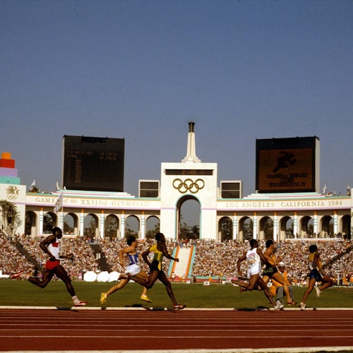 Sprinters competing in a race at the 1984 Los Angeles Olympics, with a packed stadium and Olympic rings visible in the background.