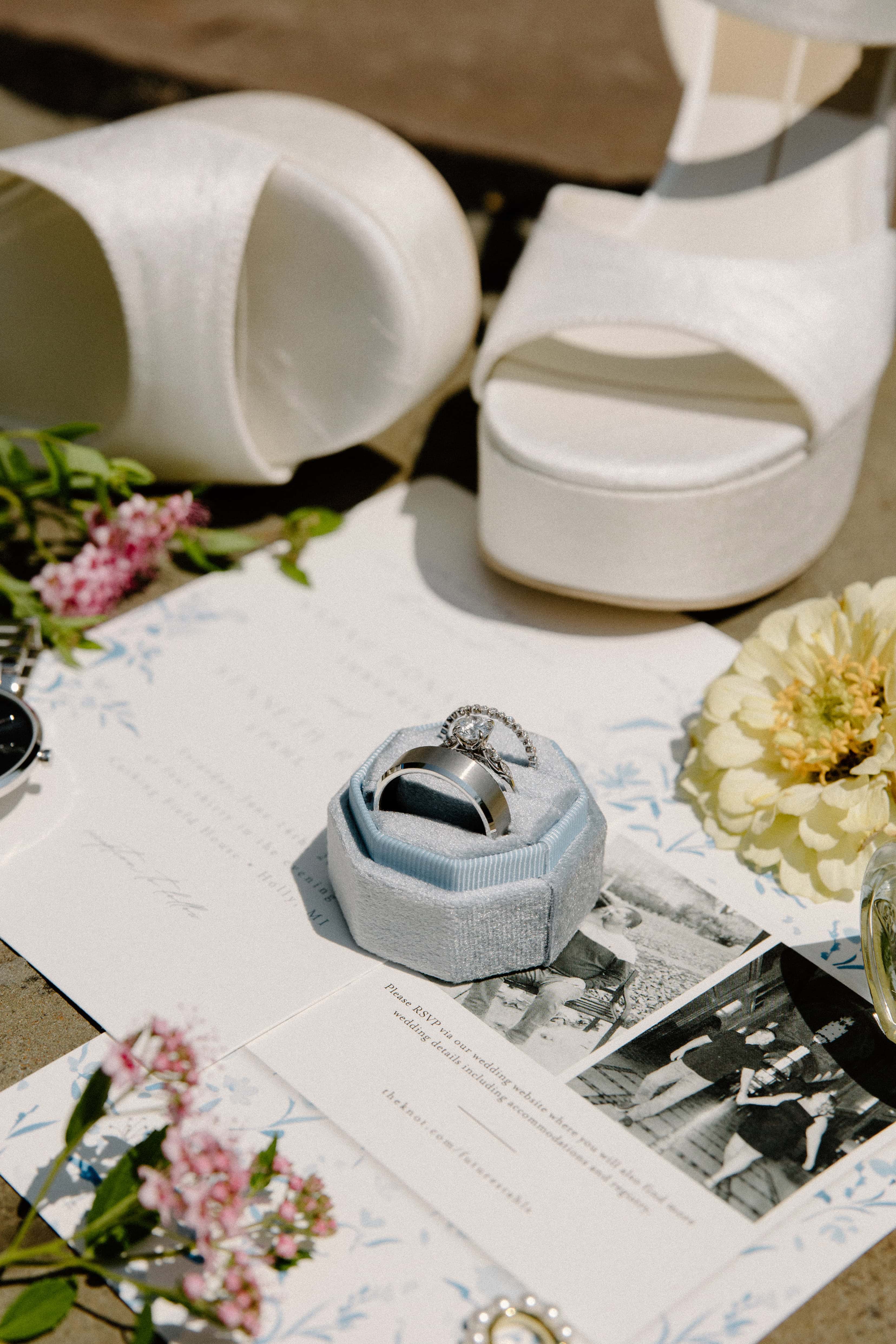 A close-up, flat lay photo of wedding accessories, including a white high-heeled shoe, a ring box with a ring inside, and a paper with text and floral illustrations on a wooden table.